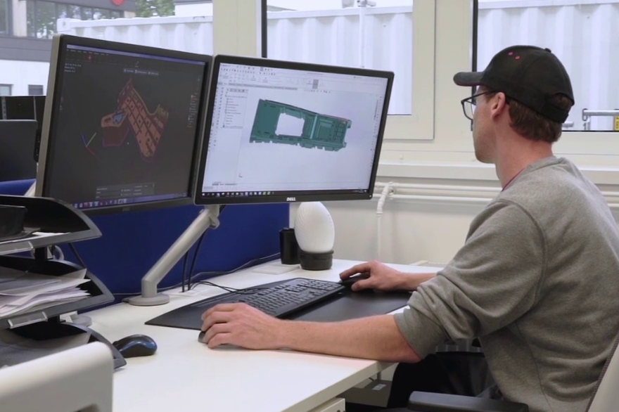 Hombre con suéter gris y gorra negra observando un diseño CAD en la pantalla de un ordenador.